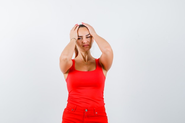 Jeune femme en débardeur rouge, pantalon tenant la tête avec les mains et à la colère, vue de face.