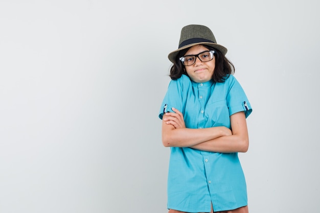 Jeune femme debout avec les bras croisés en chemise bleue, chapeau, lunettes et à la stupide