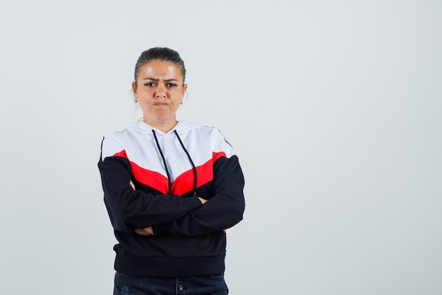 Jeune femme debout avec les bras croisés tout en acidifiant son visage en sweat-shirt coloré et à la vue offensée, de face.