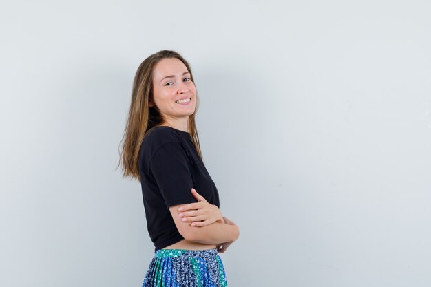 Jeune femme debout avec les bras croisés tout en posant à la caméra en t-shirt noir et jupe bleue et à la recherche de plaisir