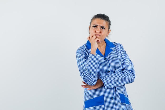 Jeune femme debout dans la pensée pose en chemise de pyjama vichy bleu et regardant pensif, vue de face.