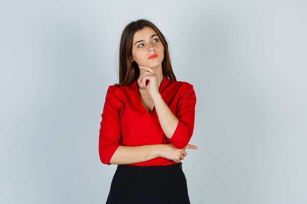 Jeune femme debout dans la pensée pose en chemisier rouge, jupe et à la pensif