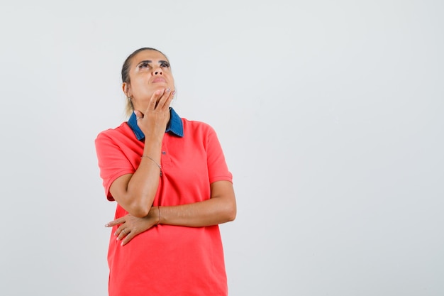 Jeune femme debout dans la pensée pose en t-shirt rouge et regardant pensif, vue de face.