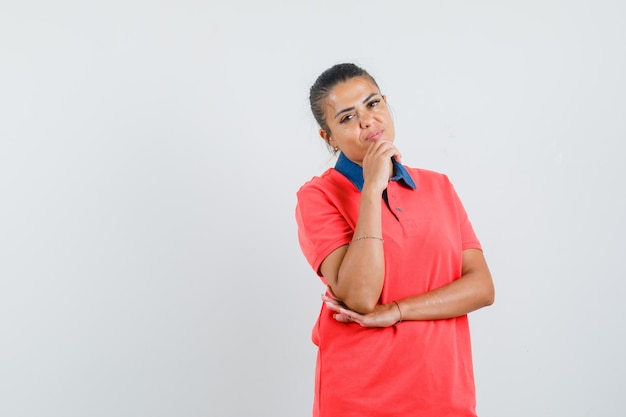 Jeune femme debout dans la pensée pose tout en mettant la main sous le menton en chemise rouge et à la vue pensive, de face.