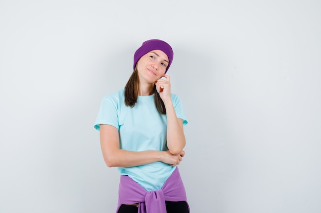 Jeune femme debout dans une pose de réflexion, la joue appuyée sur la main en t-shirt bleu, bonnet violet et l'air pensif, vue de face.