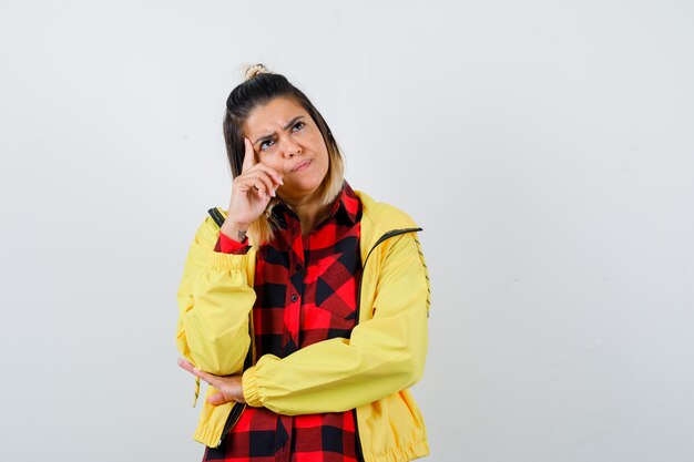 Jeune femme debout dans une pose de réflexion, regardant loin en chemise à carreaux, veste et l'air perplexe, vue de face.