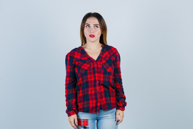 Jeune femme debout dans une pose de réflexion, regardant vers le haut en chemise à carreaux, jeans et regardant pensive. vue de face.