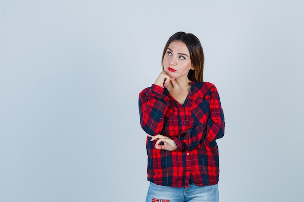 Photo gratuite jeune femme debout dans une pose de réflexion, soutenant le menton en main dans une chemise à carreaux, un jean et l'air pensif. vue de face.