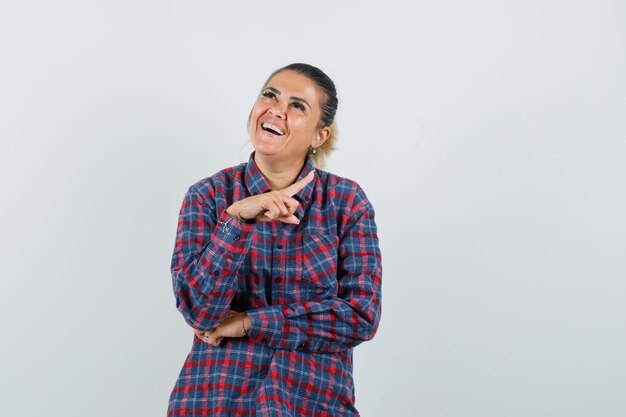 Jeune femme debout dans la pose de thinkin et pointant vers la droite avec les index en chemise à carreaux et à la recherche de plaisir. vue de face.