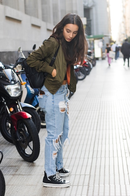 Jeune femme debout dans la rue avec un sac à dos sur l&#39;épaule en regardant quelque chose dans la poche de la veste