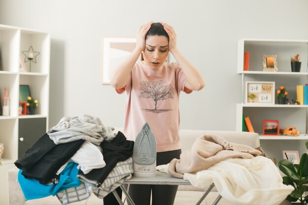 Jeune femme debout derrière une planche à repasser avec des vêtements dans le salon