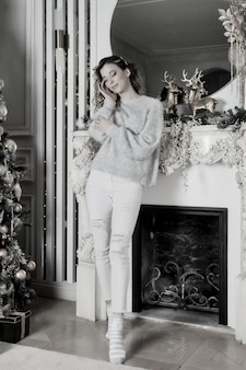 Jeune femme debout devant la cheminée et l'arbre de noël avec des cadeaux heureux et rêvant. couleur argent blanc sur fond de salon pour joyeux noël et bonne année. moments d'ambiance en famille