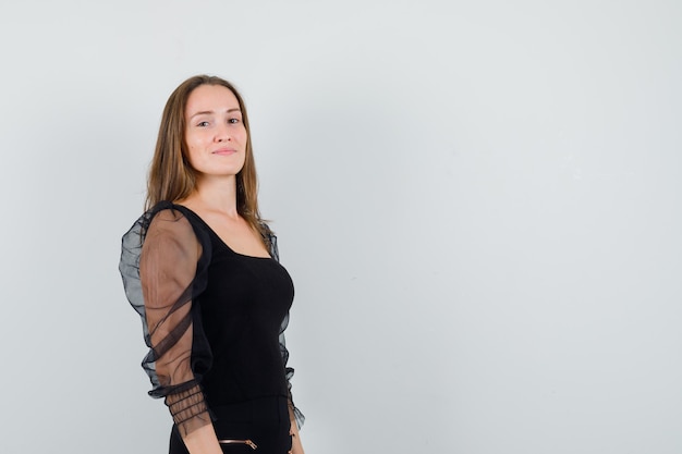 Jeune femme debout droite et posant à la caméra en chemisier noir et pantalon noir et à l'optimiste. vue de face.