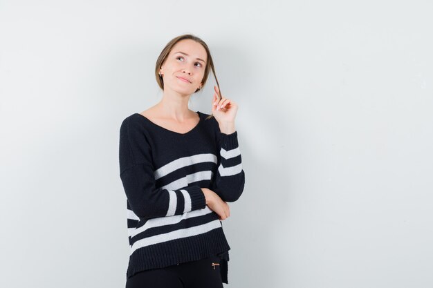 Jeune femme debout en pensant poser tout en rentrant les cheveux en maille rayée et pantalon noir et à la pensif