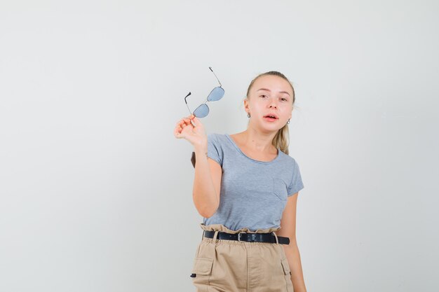 Jeune femme décollant des lunettes en t-shirt, pantalon et à la vue de face, confiant.