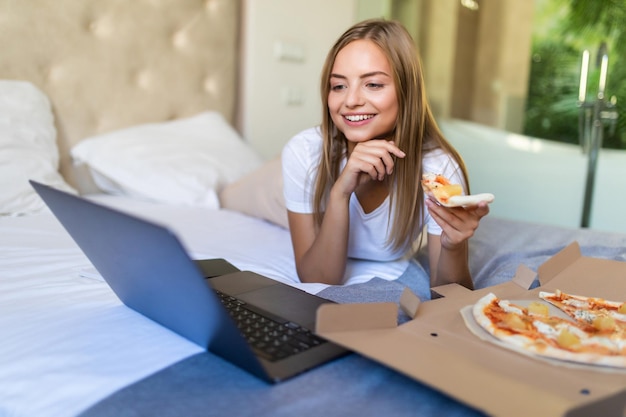 Jeune femme décontractée allongée sur un lit dans son lit à la maison tout en mangeant de la pizza et en utilisant un ordinateur portable