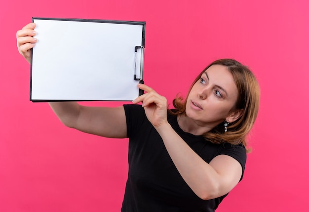 Jeune femme décontractée soulevant le presse-papiers et en le regardant sur un mur rose isolé