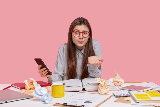 Une jeune femme désemparée hésitante hausse les épaules, regarde la caméra avec confusion, tient un téléphone portable moderne