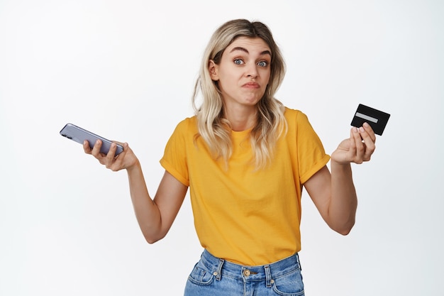 Une jeune femme désemparée tient un smartphone et une carte de crédit, haussant les épaules avec une expression de visage perplexe sur blanc.