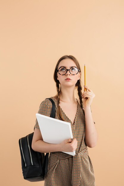 Jeune femme avec deux tresses en combinaison en tweed et lunettes modernes avec sac à dos noir sur l'épaule tenant un livre à la main et un crayon près de la tête tout en regardant pensivement sur fond beige