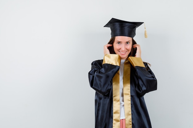 Jeune femme diplômée main dans la main près du visage en robe académique et à la recherche attrayante, vue de face.