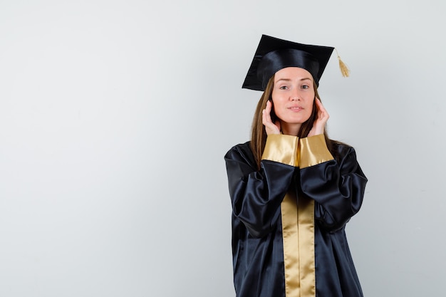 Jeune femme diplômée en robe académique, main dans la main sur les joues et à la vue de face, confiant.