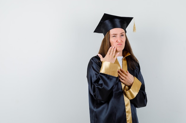 Jeune femme diplômée tenant la main sur la joue tout en gardant la main sur la poitrine isolée