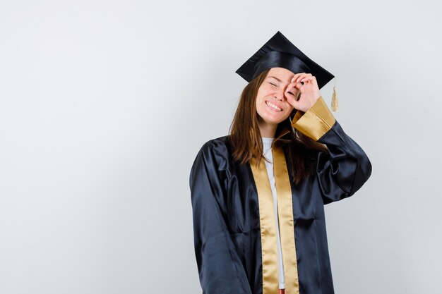 Jeune femme diplômée en tenue académique regardant à travers les doigts et à la jolie vue de face.