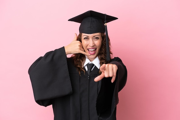Jeune femme diplômée universitaire isolée sur fond rose faisant un geste de téléphone et pointant vers l'avant
