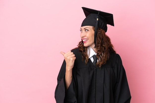 Jeune femme diplômée universitaire isolée sur fond rose pointant vers le côté pour présenter un produit