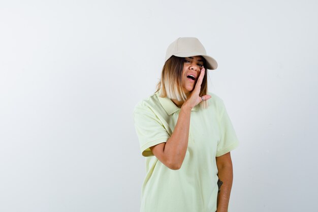 Jeune femme disant secret derrière la main en t-shirt, casquette et à la vue sérieuse, de face.