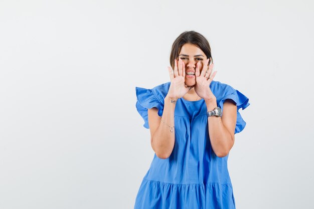 Jeune femme disant secret avec les mains près de la bouche en robe bleue et à la joyeuse