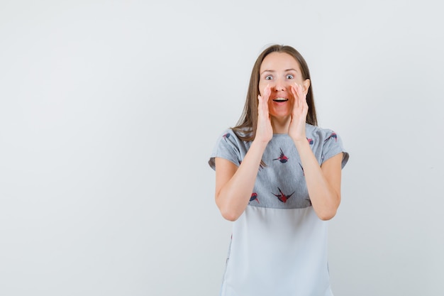 Jeune femme disant secret en t-shirt et à la curieuse. vue de face.