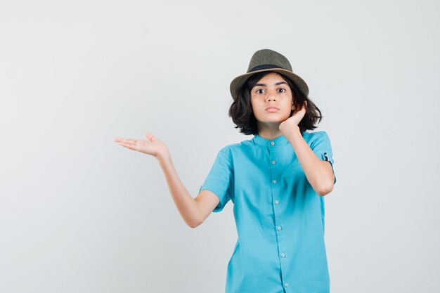 Jeune femme écartant sa paume ouverte de côté en chemise bleue, chapeau et à la surprise