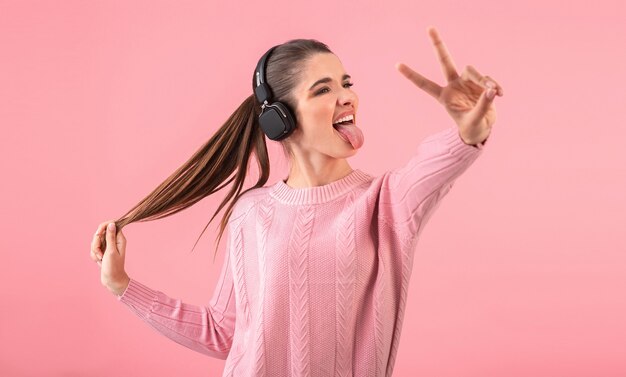 Jeune femme écoutant de la musique dans des écouteurs sans fil portant un pull rose souriant posant sur rose