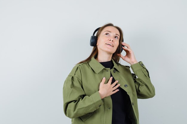 Jeune femme écoutant de la musique avec des écouteurs en veste verte et l'air satisfait, vue de face.