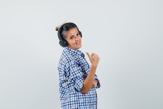 Jeune femme écoutant de la musique via des écouteurs tout en montrant le pouce en chemise à carreaux et à la joie. .