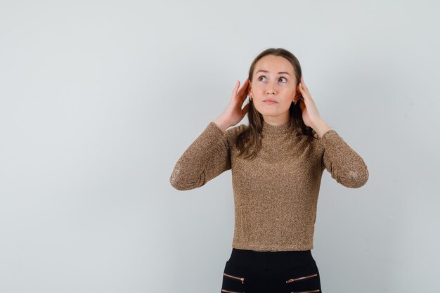 Jeune femme écoutant quelque chose tout en regardant ailleurs en chemisier doré et à la vue attentive, de face. espace pour le texte