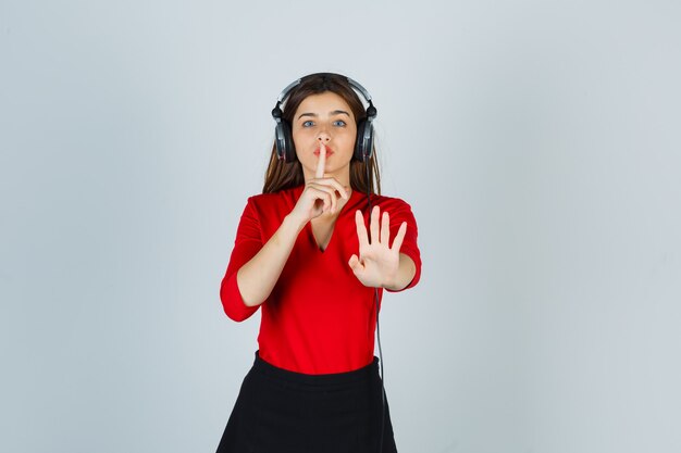 Jeune femme avec des écouteurs montrant le geste de silence en chemisier rouge