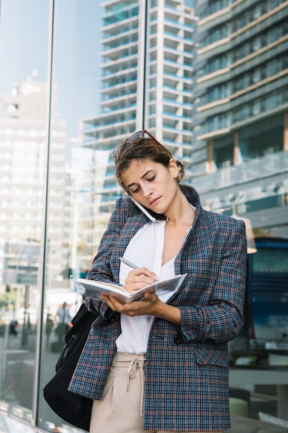 Photo gratuite jeune femme écrivant des notes dans le cahier avec un stylo contre du verre réfléchissant