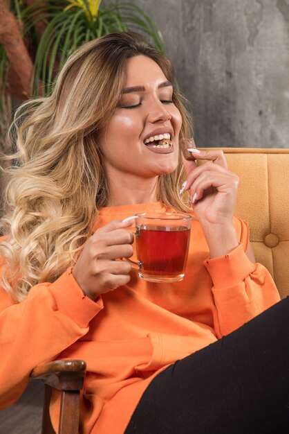 Jeune femme élégante assise dans un fauteuil, manger du chocolat et tenant du thé.