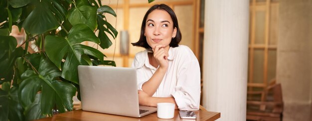 Jeune femme élégante et confiante avec un ordinateur portable assise dans un café et travaillant en indépendant dans un espace de coworking