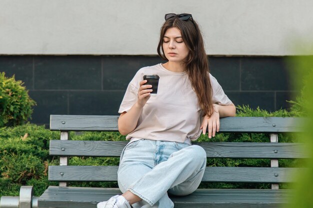 Une jeune femme élégante en lunettes de soleil est assise sur un banc et boit du café