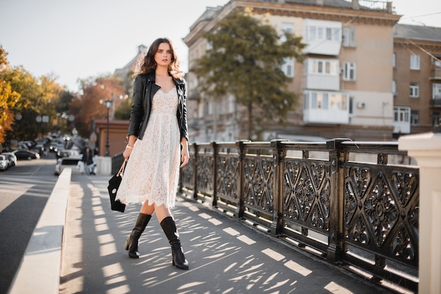 Jeune femme élégante marchant dans la rue en tenue à la mode, tenant un sac à main, portant une veste en cuir noir et une robe en dentelle blanche, style printemps automne