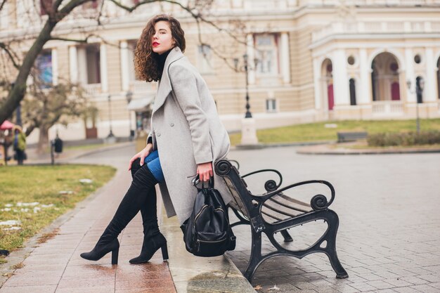 Jeune femme élégante marchant dans la ville d'automne, saison froide, portant des bottes noires à talons hauts, sac à dos en cuir, accessoires, manteau gris, assis sur un banc, tendance de la mode