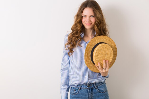 Jeune femme élégante mignonne en tenue de style été posant sur un mur blanc tenant un chapeau de paille