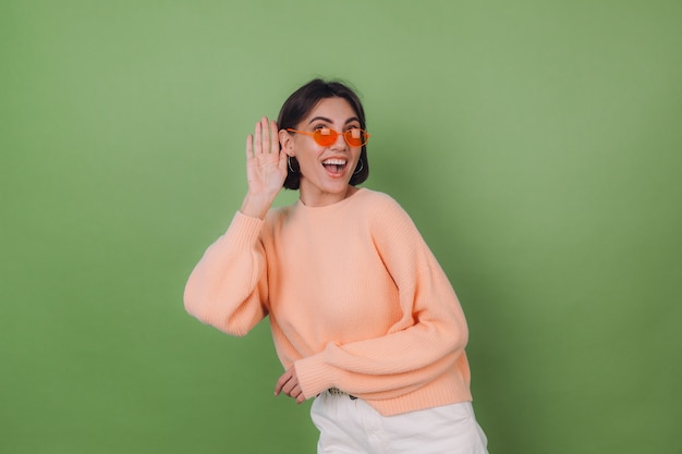 Jeune femme élégante en pull pêche occasionnel et lunettes orange isolé sur mur d'olive verte curieux essayez d'entendre ce que vous dites avec la main par l'oreille copie espace