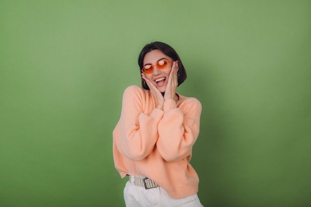 Jeune femme élégante en pull pêche occasionnel et lunettes orange isolé sur mur d'olive verte heureux rire souriant positif autour de l'espace de copie