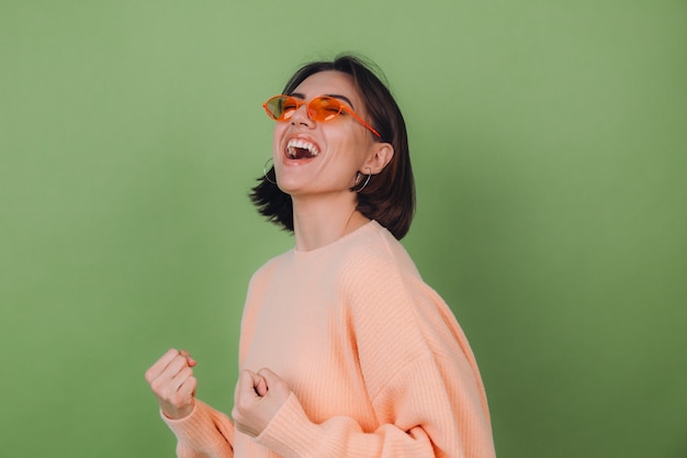 Jeune femme élégante en pull pêche occasionnel et lunettes orange isolé sur mur d'olive verte positive faisant geste gagnant poing serrant l'espace de copie