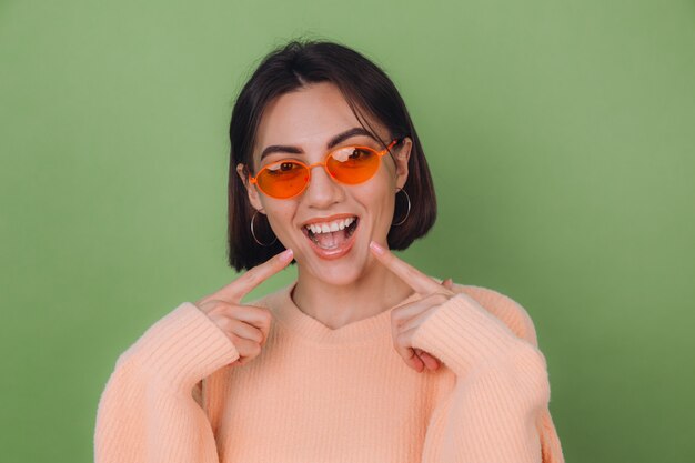 Jeune femme élégante en pull pêche occasionnel et lunettes orange isolé sur mur d'olive verte souriant positif pointant sur les dents blanches avec index copie espace